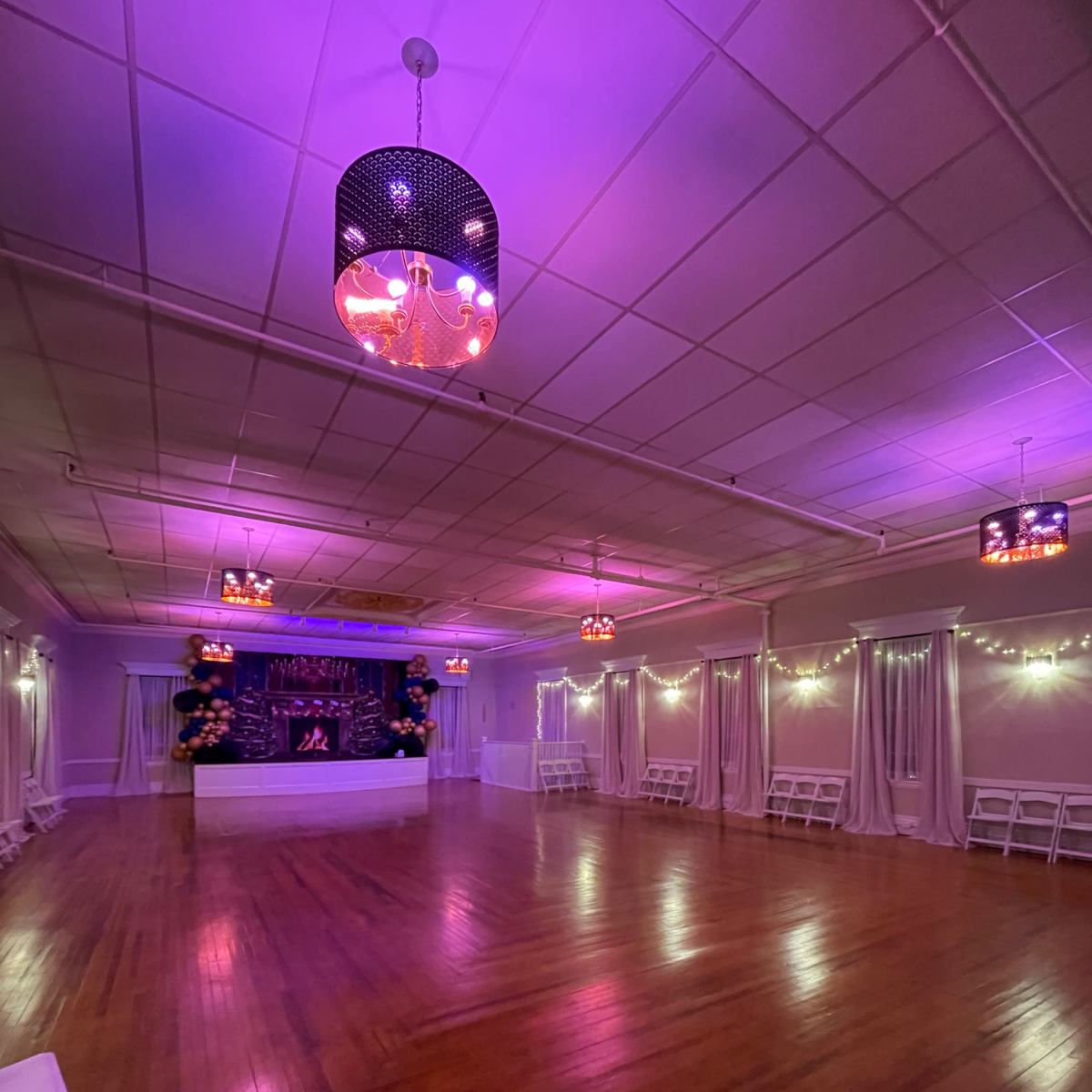 An empty event space with wooden flooring, purple lighting, and white folding chairs arranged along the sides.