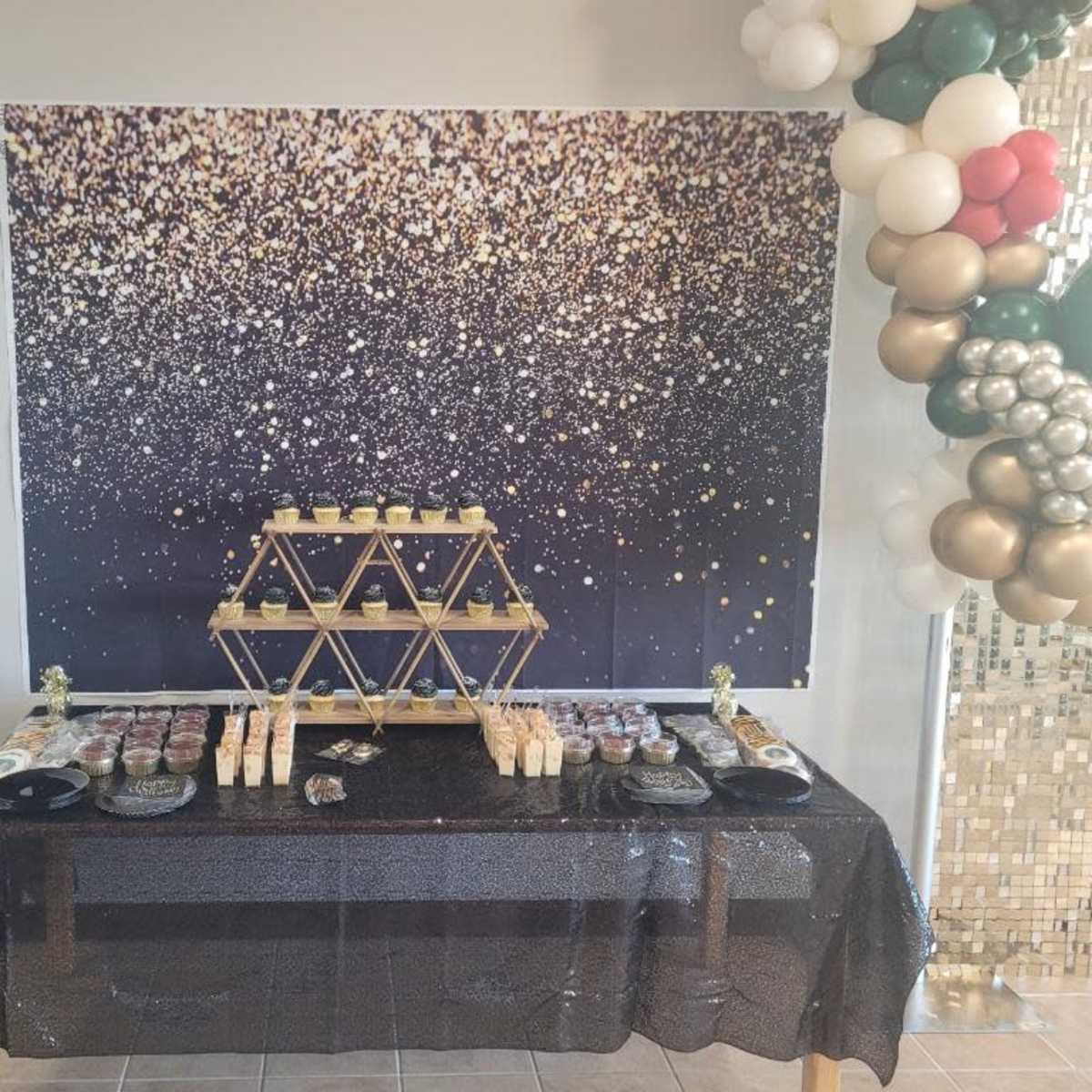 A decorated table covered with a black tablecloth, displaying an assortment of desserts, set against a sparkly gold and black backdrop.