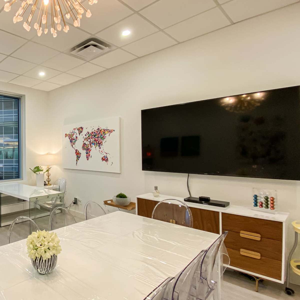 A modern conference room features a large window with a city view, a white table, transparent chairs, and a wall-mounted TV.