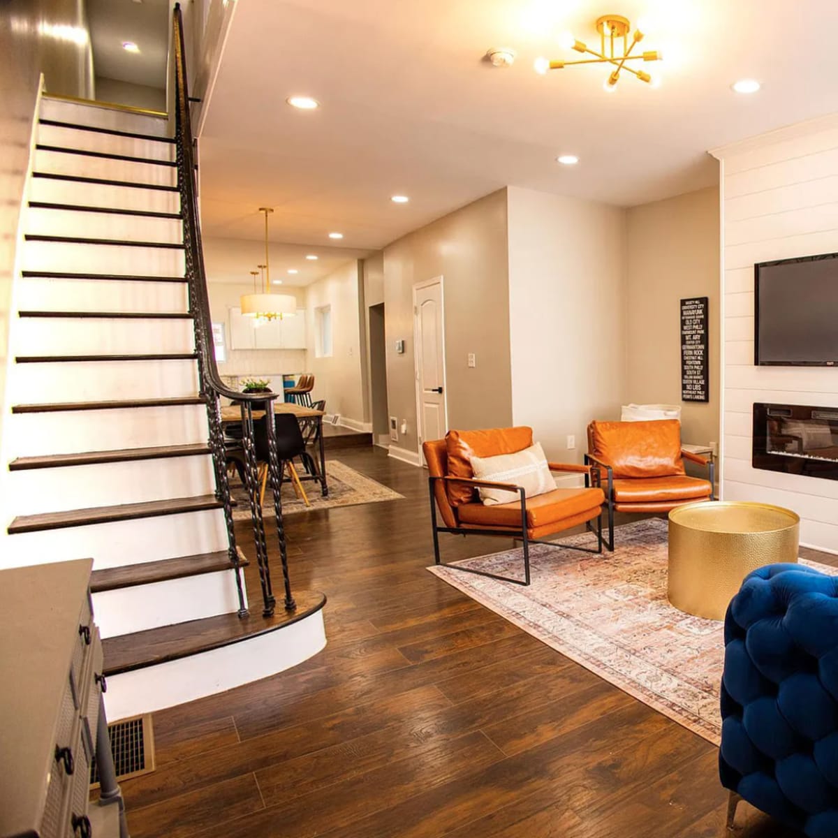 A modern living room with a blue tufted sofa, two orange armchairs, a gold coffee table, a mounted TV, and a staircase in the background.