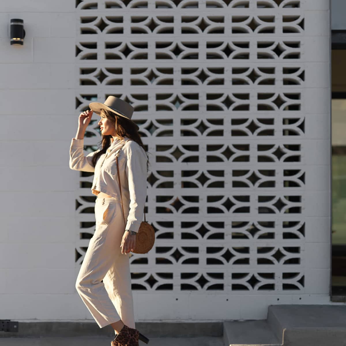A woman in a light-colored outfit and hat walks beside a textured white wall with a geometric design.