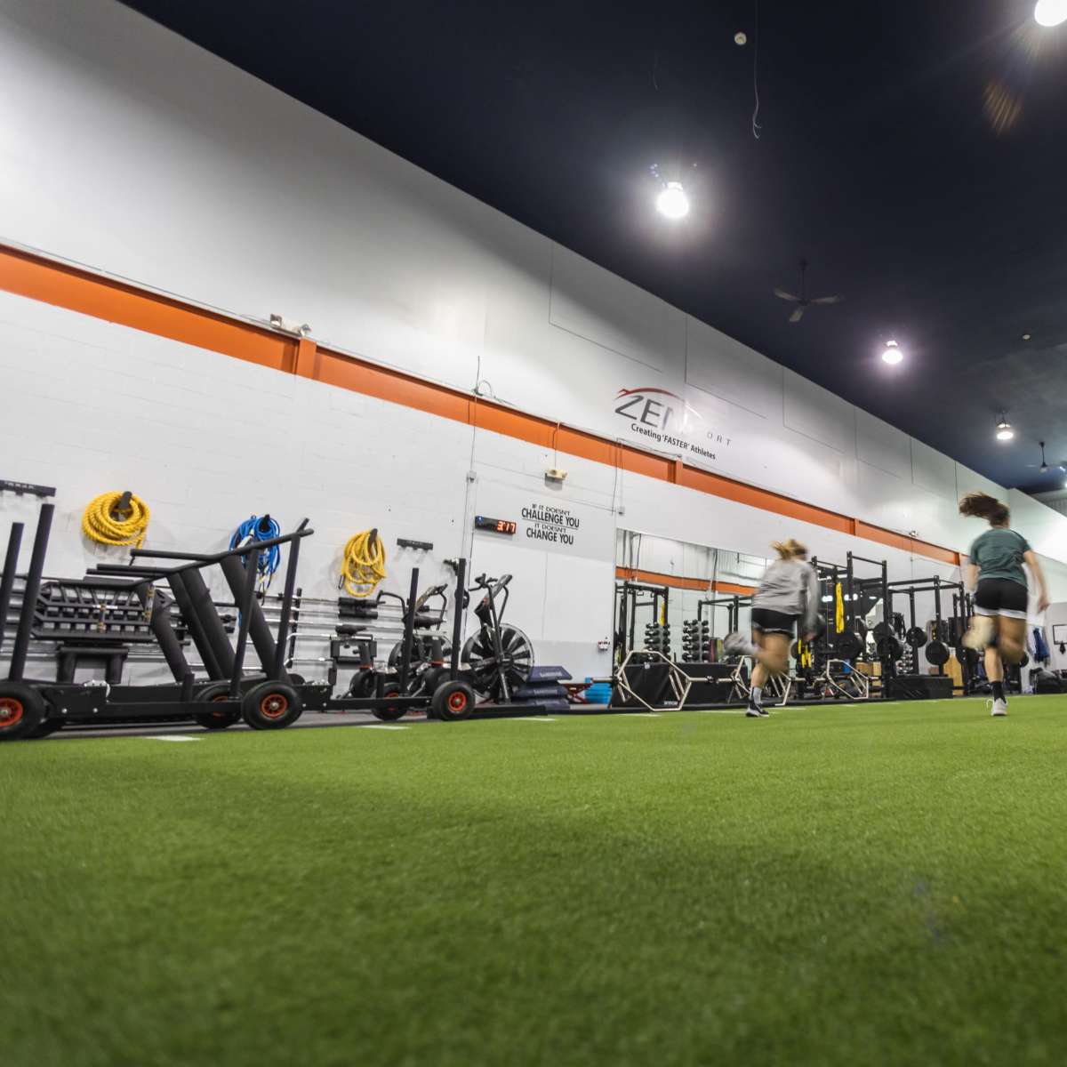 A group of four athletes runs on a turf surface inside a gym equipped with fitness equipment along the walls.