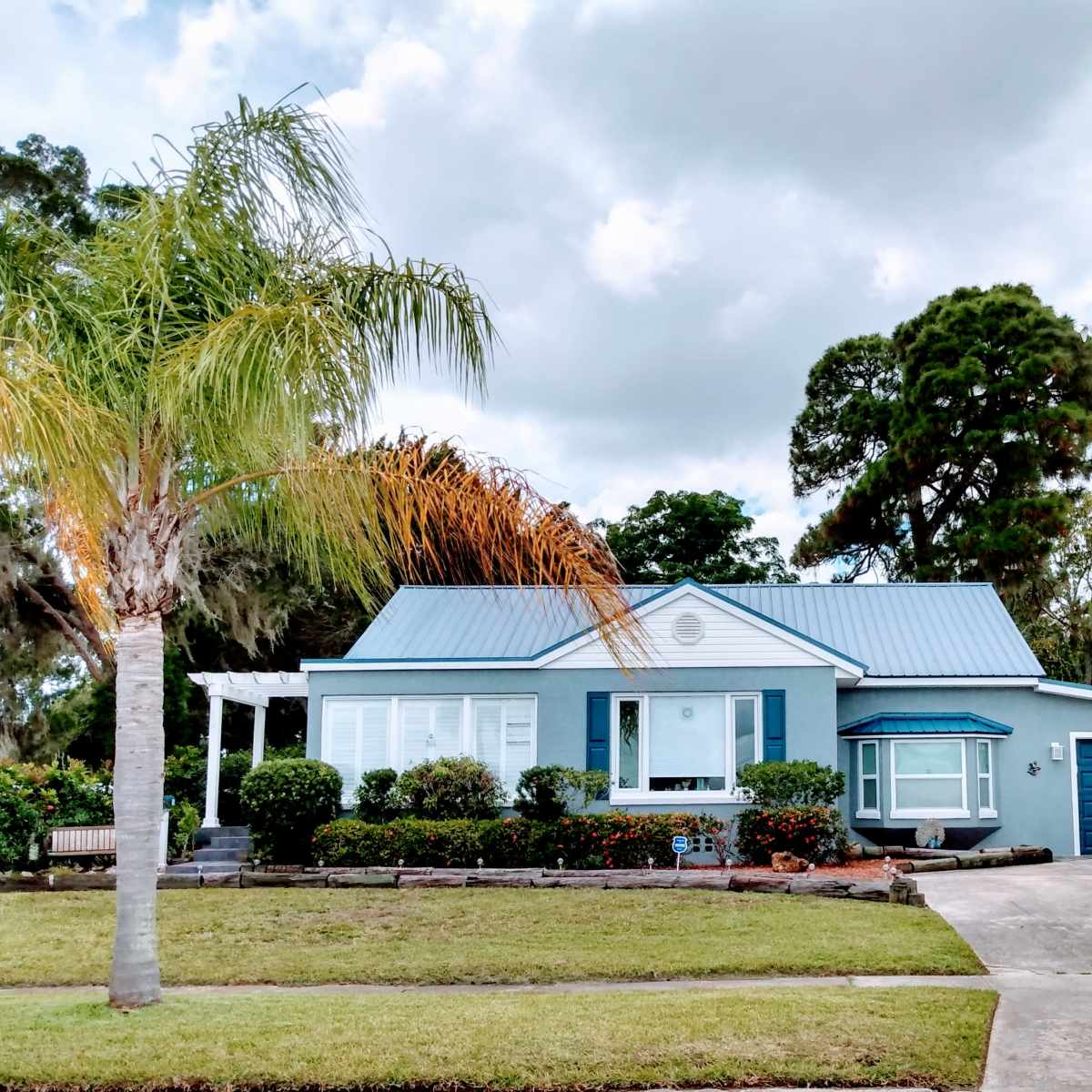A light blue house with a gray metal roof and a blue garage door is surrounded by neatly trimmed landscaping and palm trees.