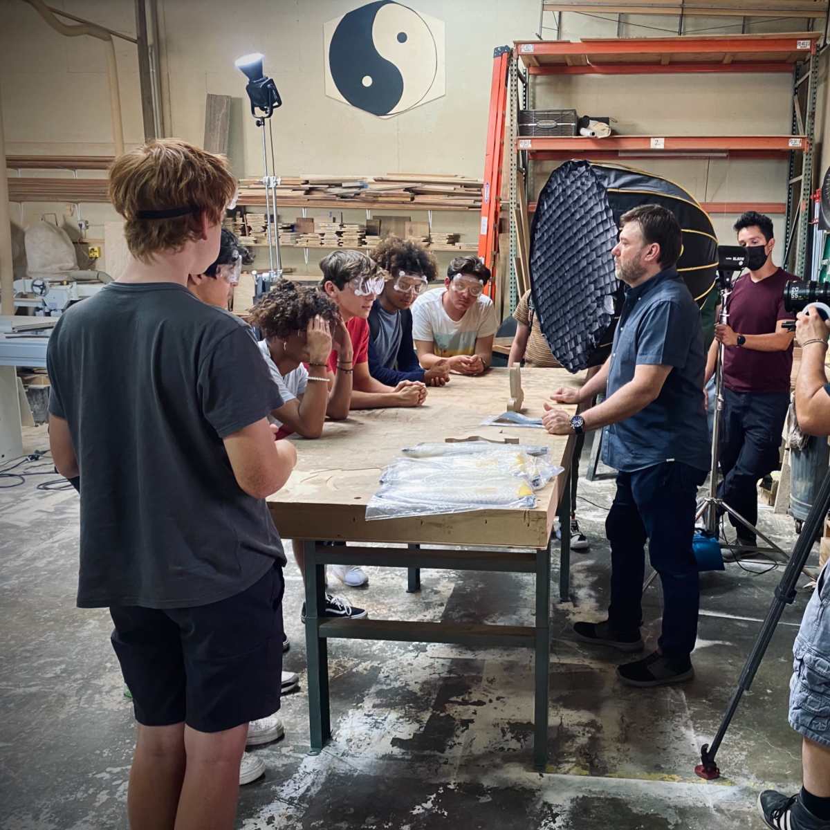 A group of people is gathered around a table in a workshop while one person speaks to them, and a camera is recording the interaction.