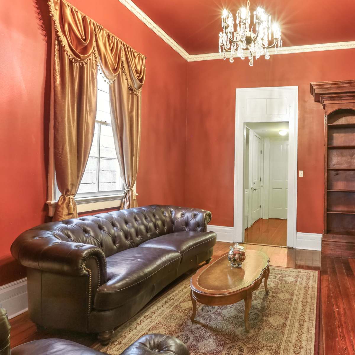 A formal living room with dark red walls, a leather sofa, a wooden bookshelf, and a decorative chandelier.