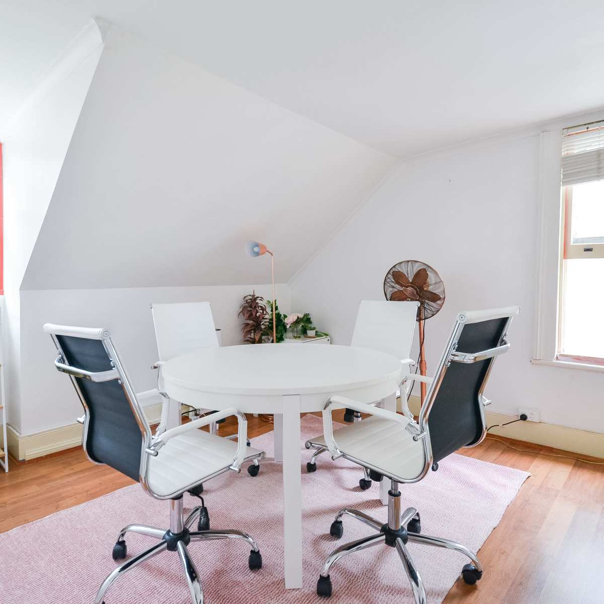 A small, well-lit room features a round white table surrounded by four black office chairs, with plants and a fan in the corners.