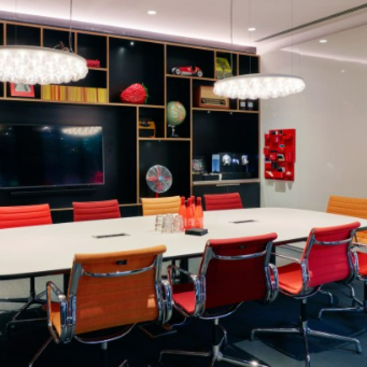 A modern conference room with a large white table surrounded by red and orange chairs, featuring decorative shelves with various items and a television mounted on the wall.