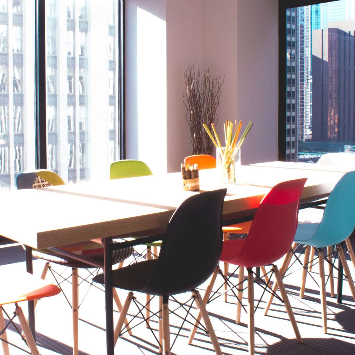 A conference room features a large table surrounded by brightly colored chairs, with large windows showing a cityscape.