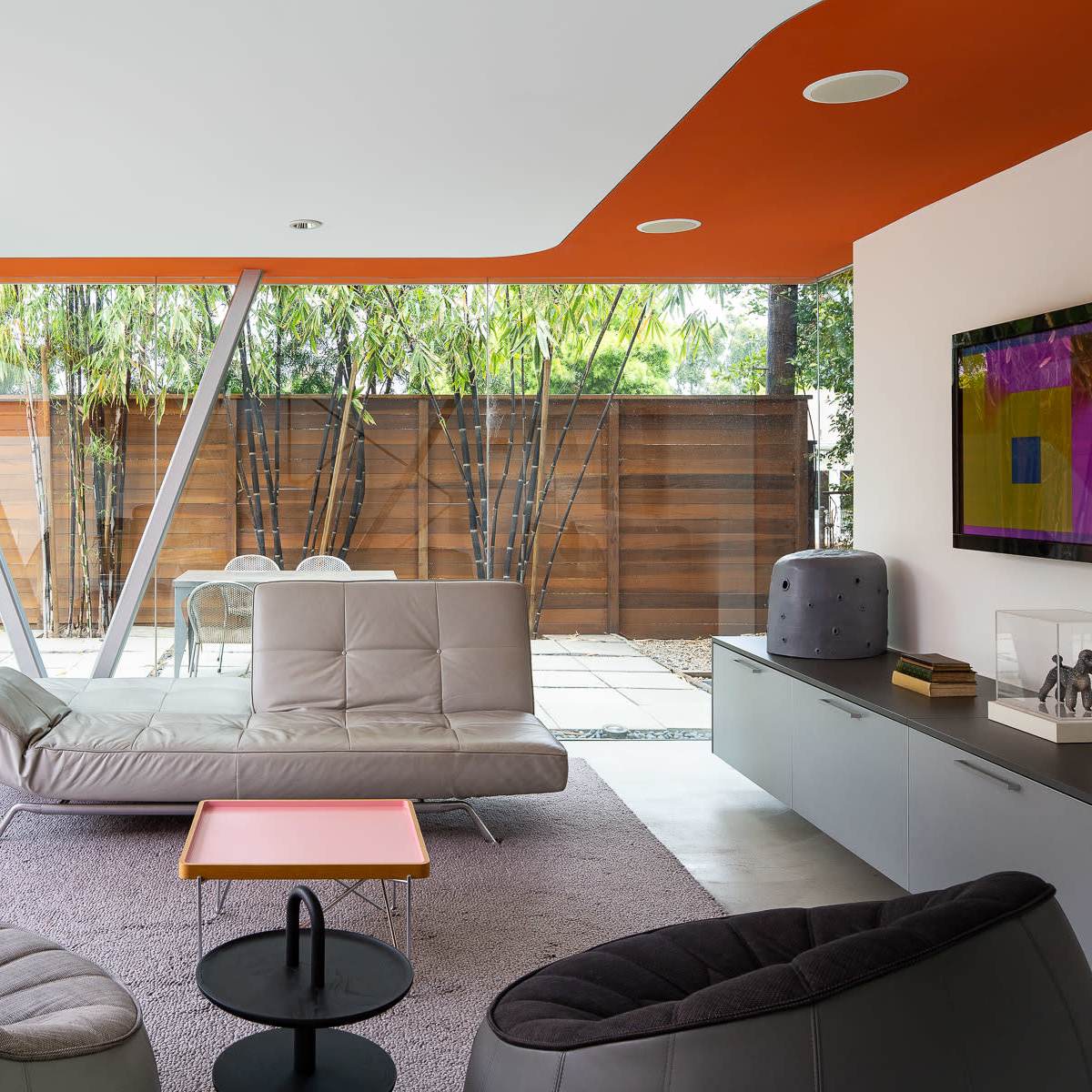 A modern living room with a gray sectional sofa, two black lounge chairs, and floor-to-ceiling windows that overlook a landscaped backyard with wooden fencing.