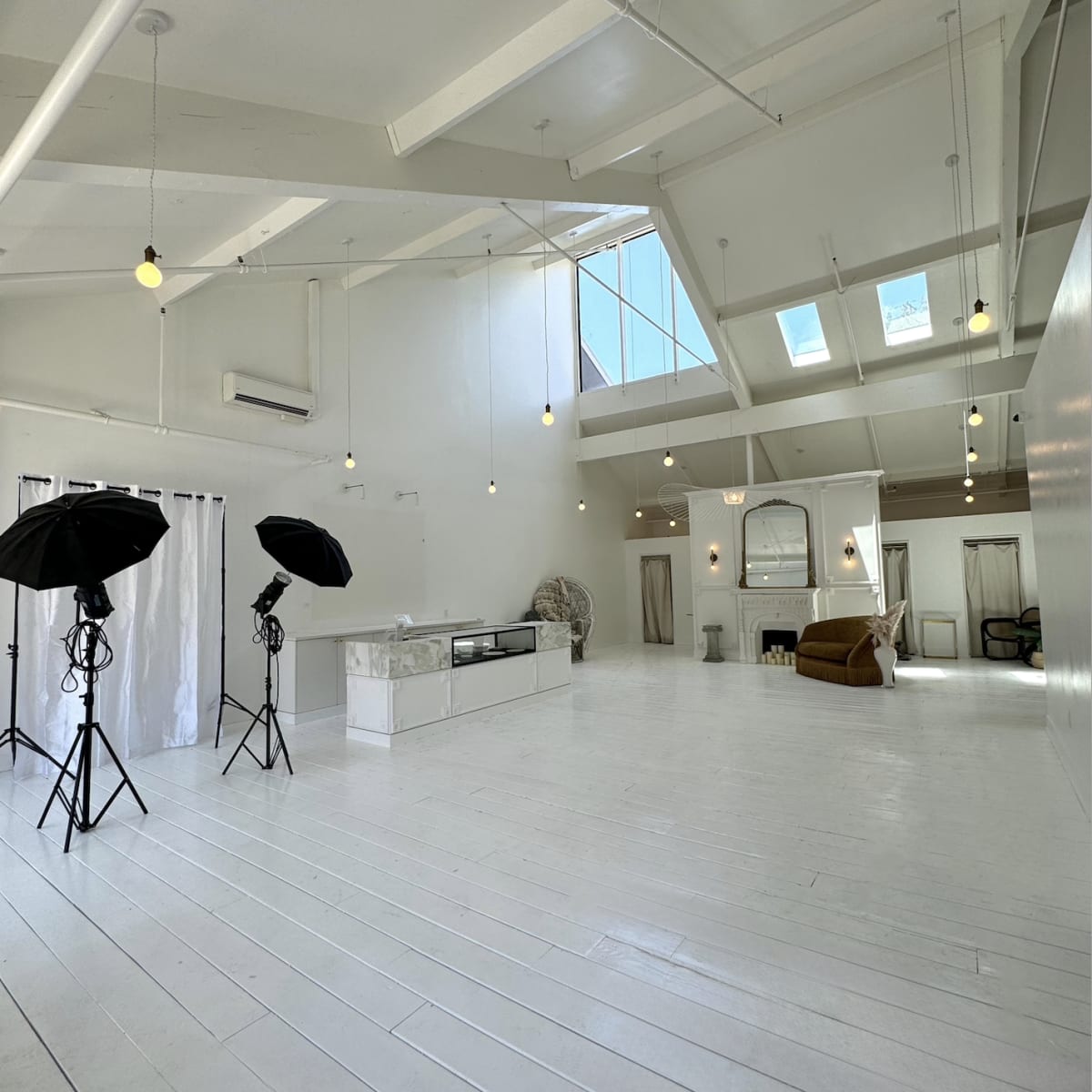 A spacious, well-lit studio featuring white wooden floors, large windows, and photography equipment set up on one side.