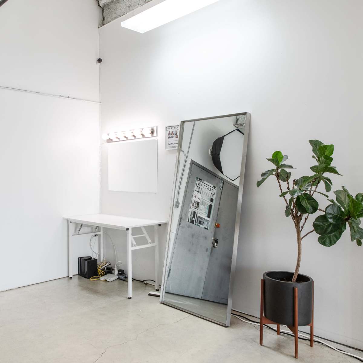 A minimalist room with a large mirror, a small table, and a potted plant next to a door with posters.
