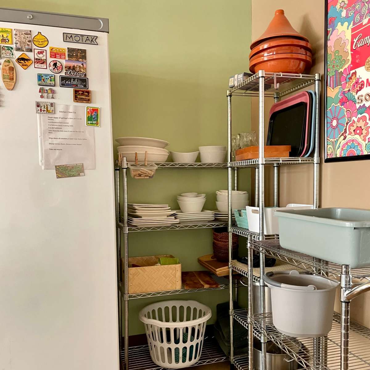 A kitchen corner with a refrigerator, shelves filled with dishes and utensils, and a laundry basket on the floor.