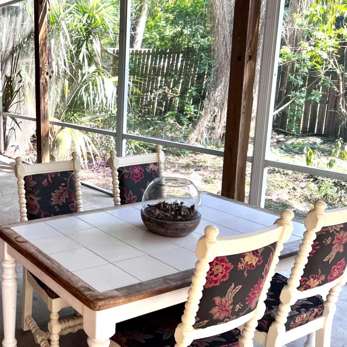 A screened-in porch features a table with four chairs and a decorative bowl on top, surrounded by greenery and a sheer curtain.