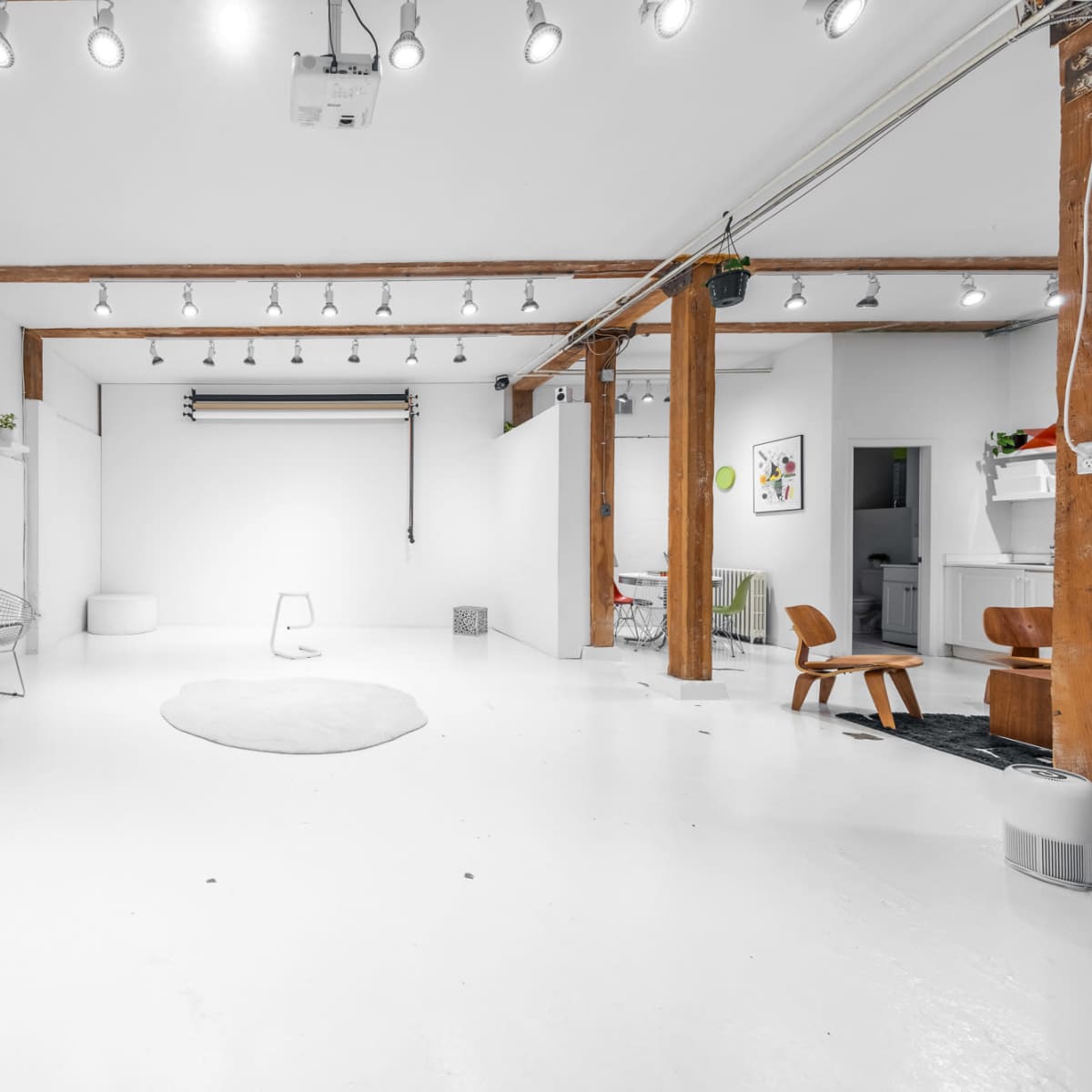 A spacious, minimalist studio with white walls, a light-colored floor, and wooden beams, furnished with a few modern chairs and a small TV unit.