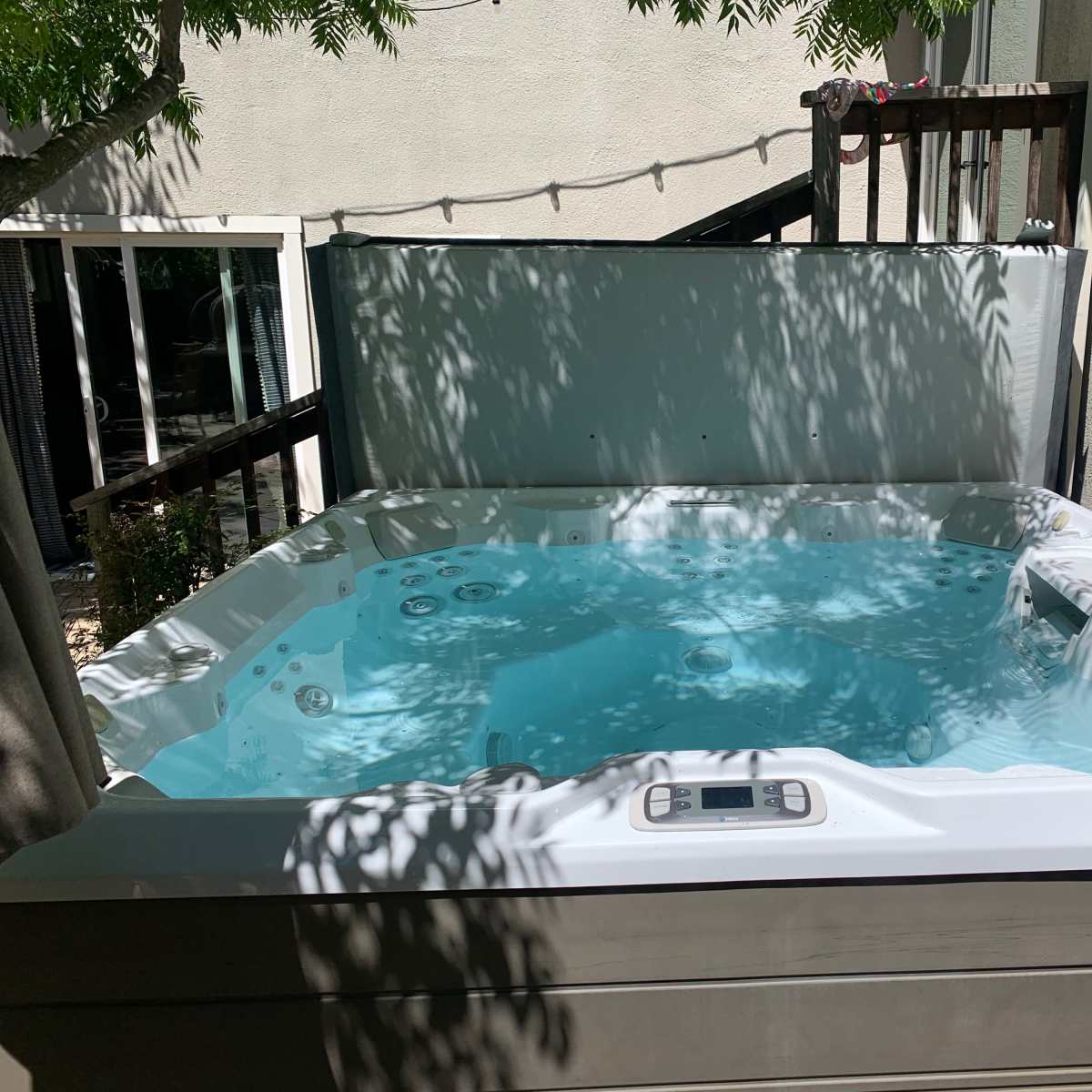 A hot tub filled with clear water is situated in a shaded area surrounded by a fence and trees.