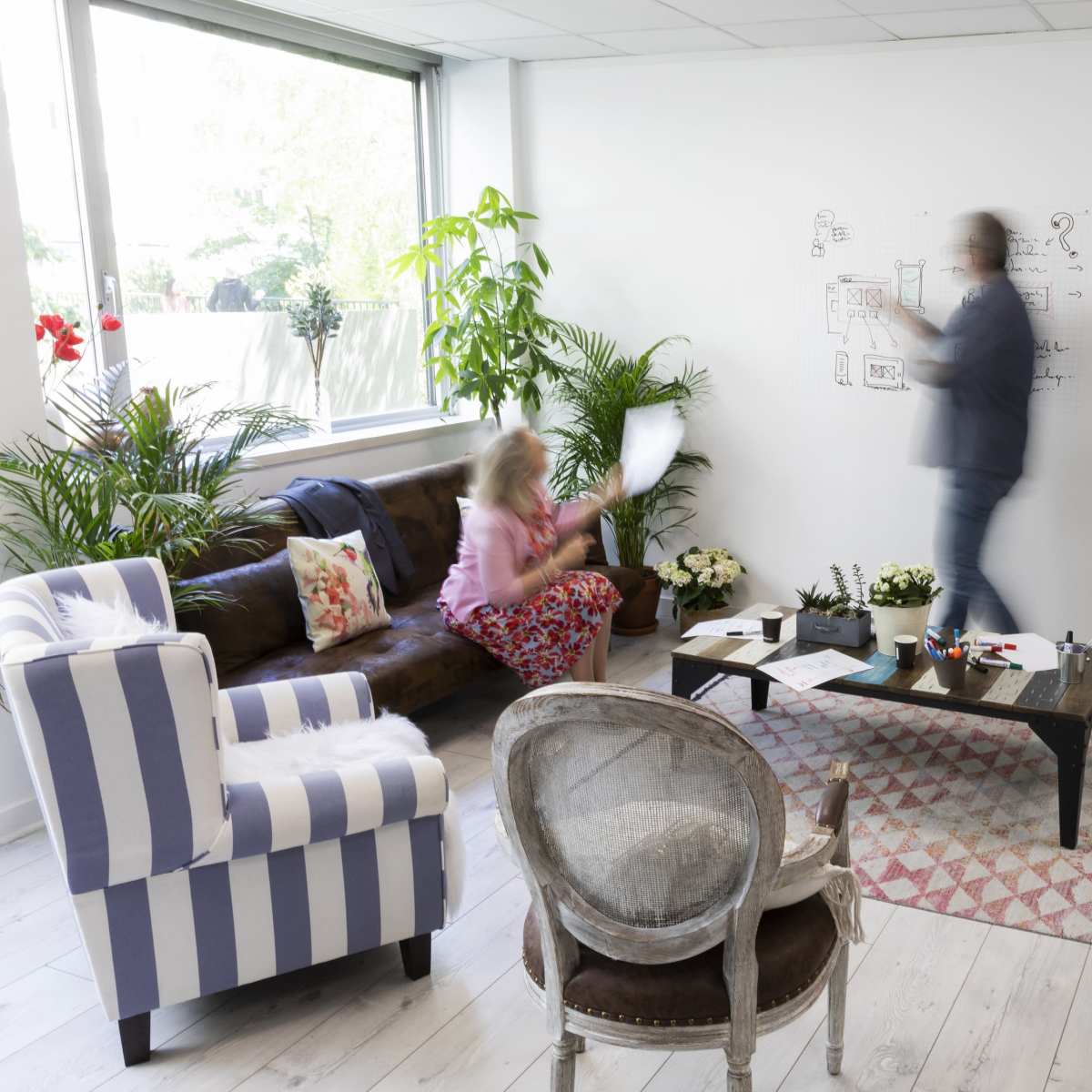 A person writes on a whiteboard while another sits on a couch holding a paper in a brightly lit, plant-filled office space.