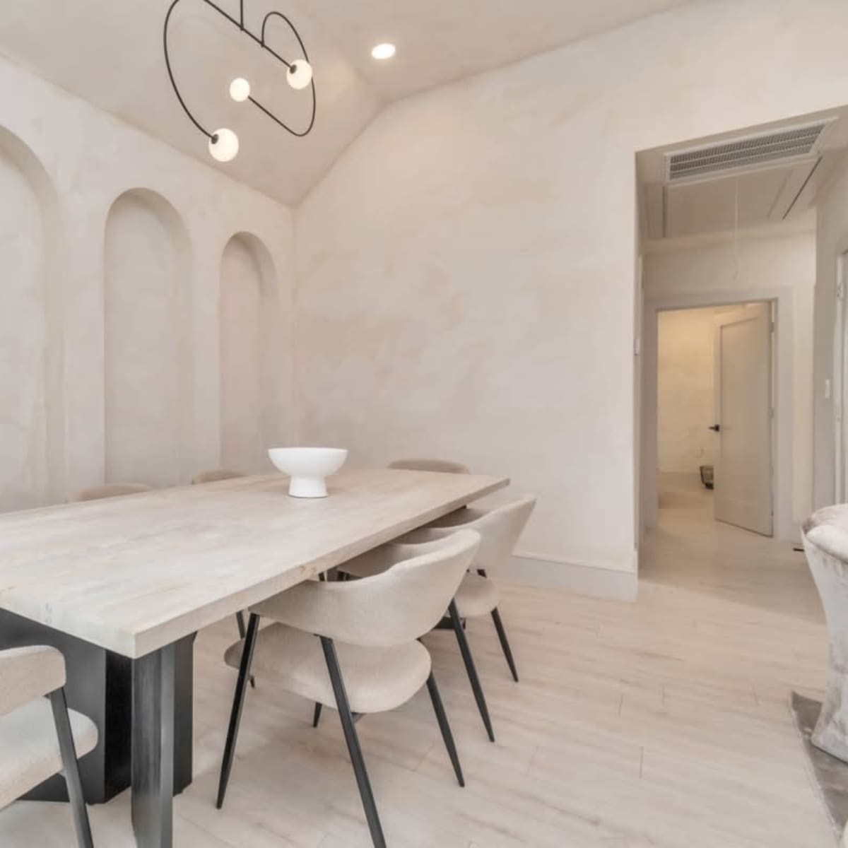 A minimalist dining room features a long wooden table surrounded by upholstered chairs beneath a modern light fixture and accented by curved wall niches.
