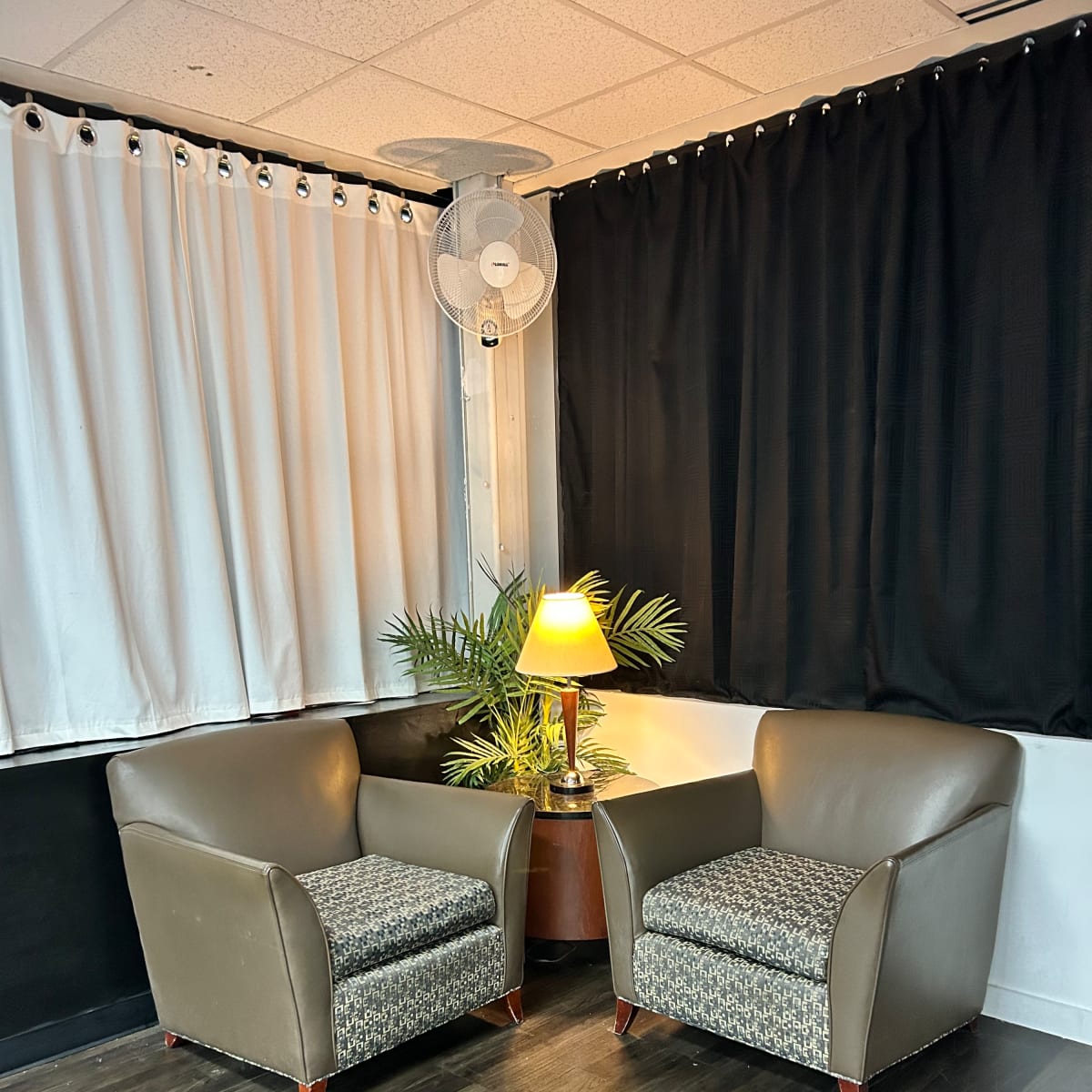 Two upholstered armchairs flank a small wooden table with a lamp and a potted plant in a room with black and white curtains.