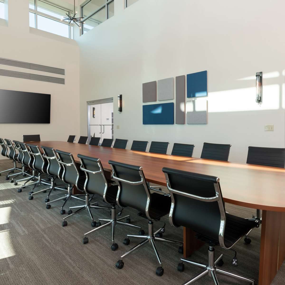A large, modern conference room features a long wooden table with black rolling chairs and a wall-mounted screen.