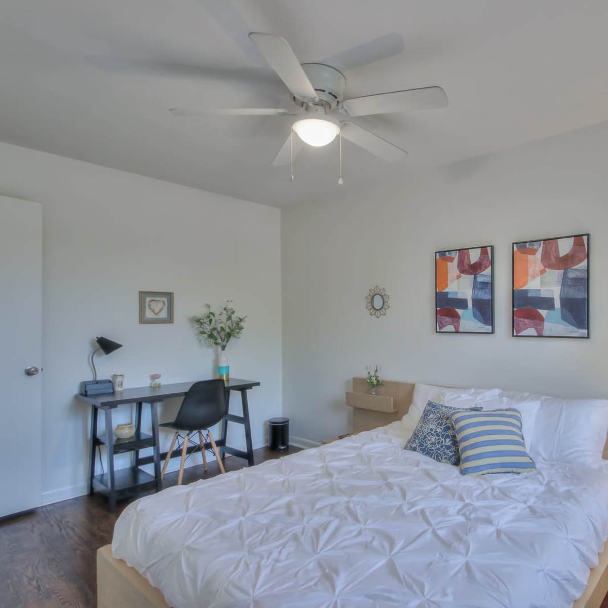 A modern bedroom featuring a bed, two decorative wall prints, a desk with a lamp, and a ceiling fan.