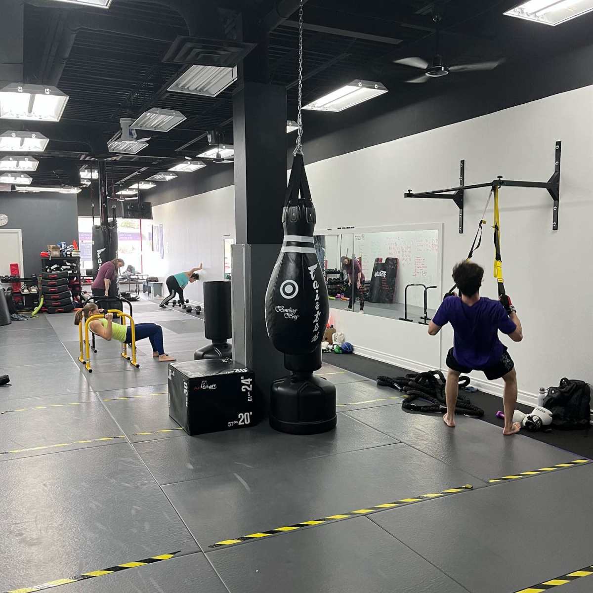 A gym interior features several workout stations, including a punching bag, TRX suspension trainer, and individuals exercising with various equipment.