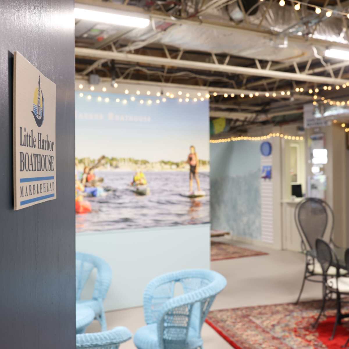 The interior of a boathouse facility with a sign for "Little Harbor Boathouse," featuring seating areas, decorative lighting, and a backdrop of people kayaking.