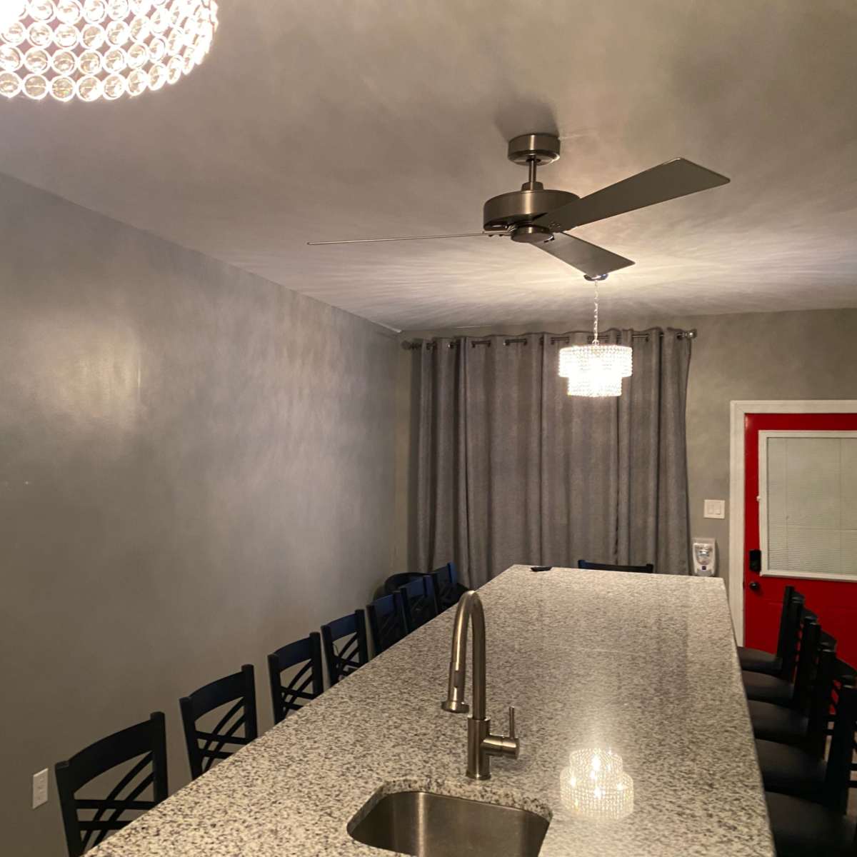A long granite countertop with black bar stools is illuminated by two chandeliers, alongside gray curtains and a red door in the background.