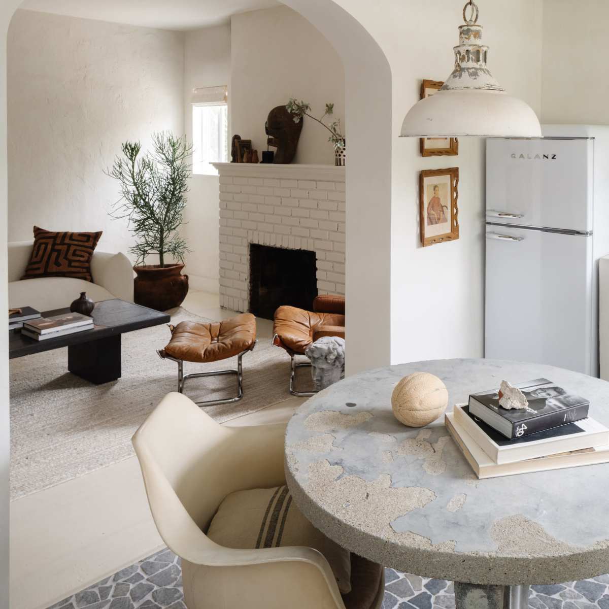 A minimalist kitchen and living area featuring a round concrete table with books on it, a retro-style refrigerator, and a cozy seating area with a fireplace.
