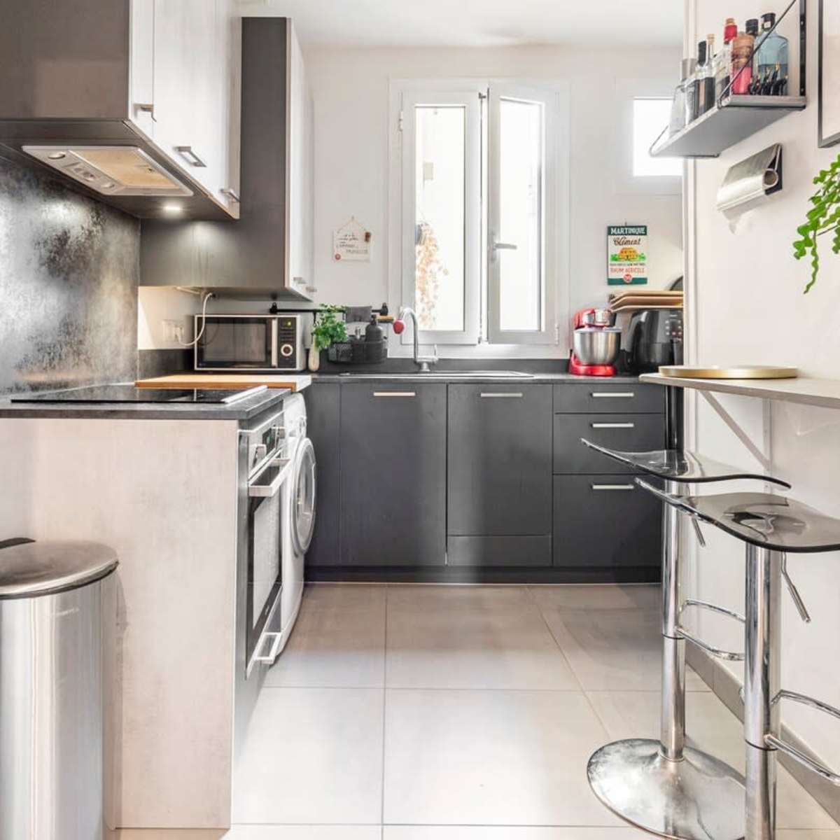 A modern kitchen with dark cabinetry, a stainless steel refrigerator, a washing machine, and a small bar table with stools.