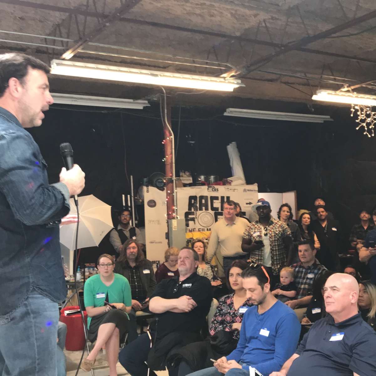 A man stands on a stage speaking into a microphone while an audience listens attentively in a warehouse setting.