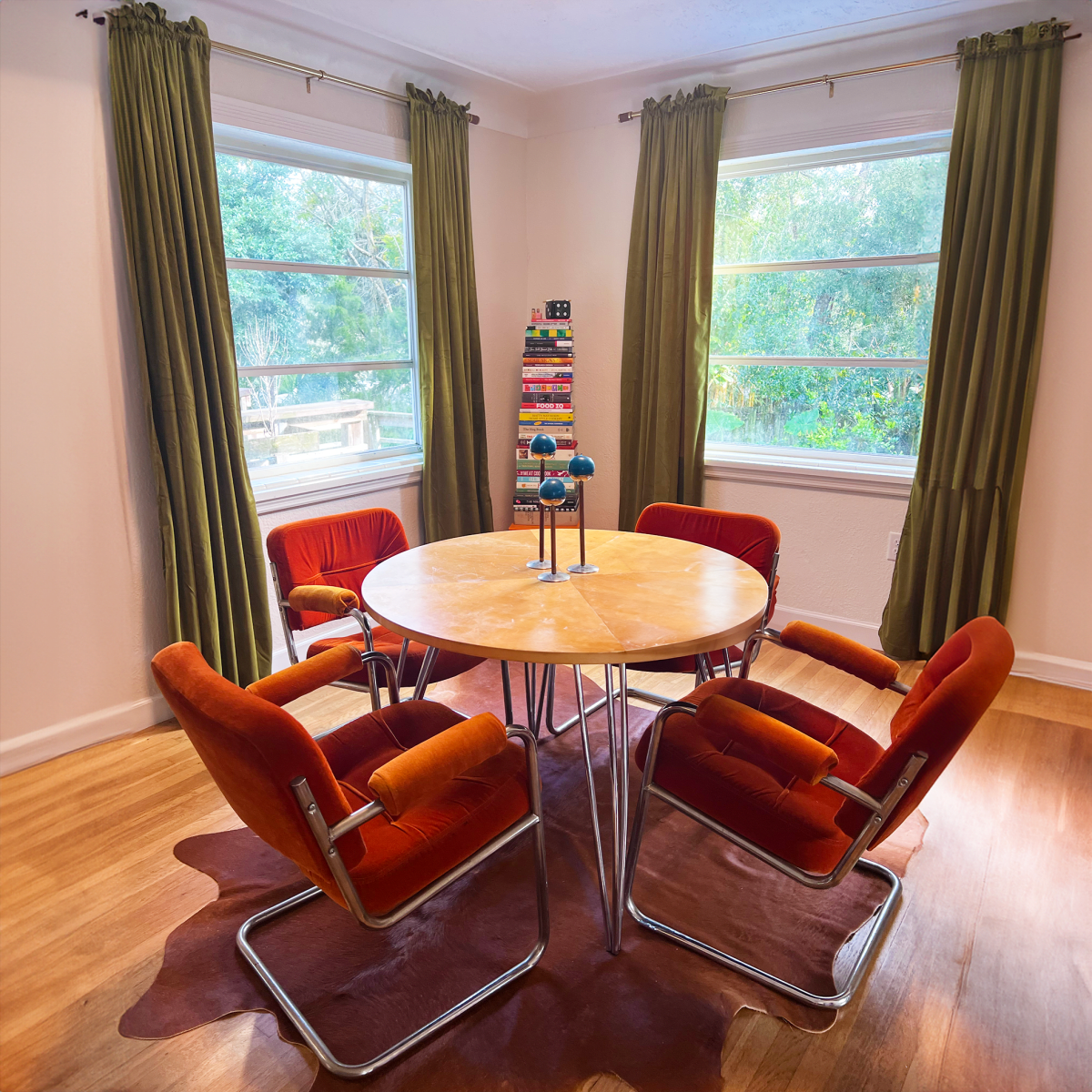 A round wooden dining table surrounded by four orange chairs is situated in a well-lit room with green curtains and large windows.