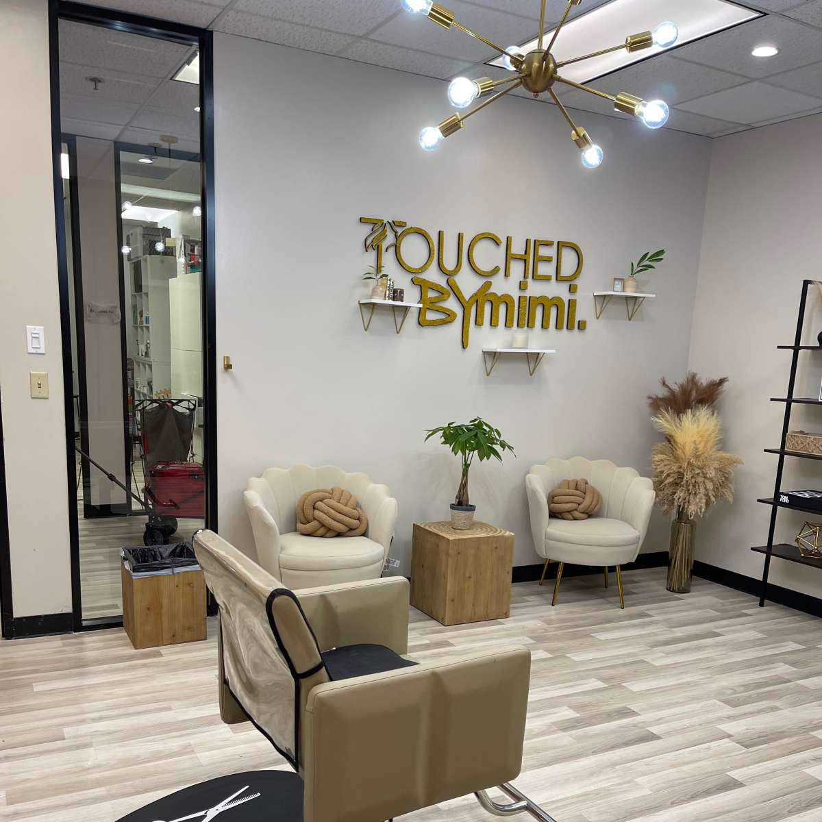 A salon interior featuring a styling chair, two armchairs, decorative plants, and a wall sign that reads "Touched By Mimi."