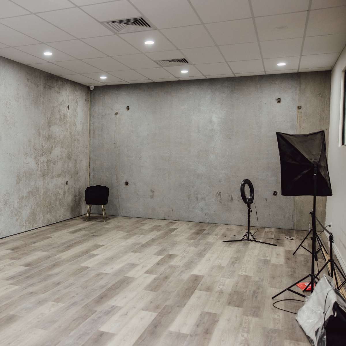 An empty room with a concrete-textured wall, wooden flooring, and minimal lighting equipment in one corner.