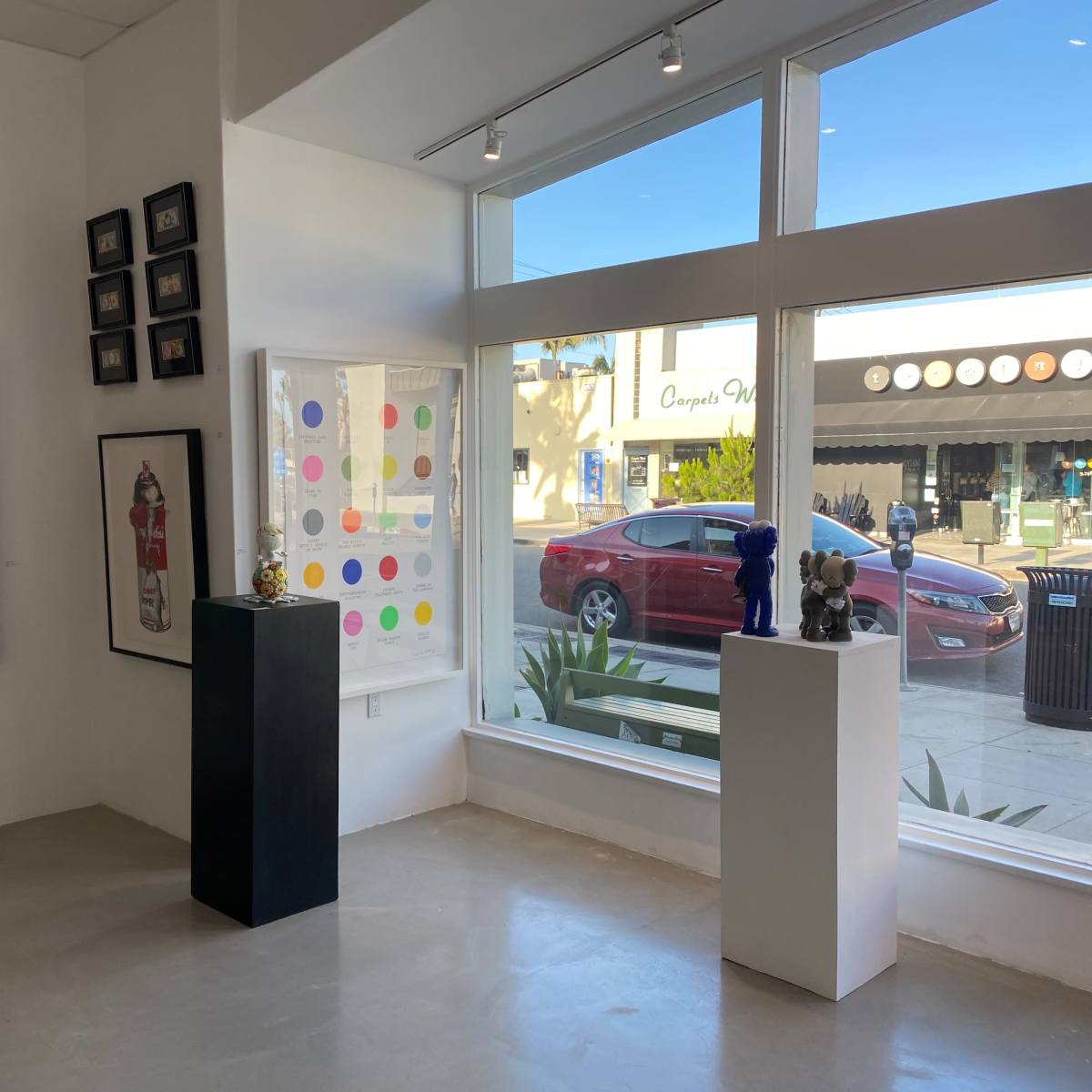 A brightly lit gallery interior featuring a large pop art-style framed print on the wall, multiple framed pictures, and sculptures displayed on white pedestals with a view of a street outside through large windows.