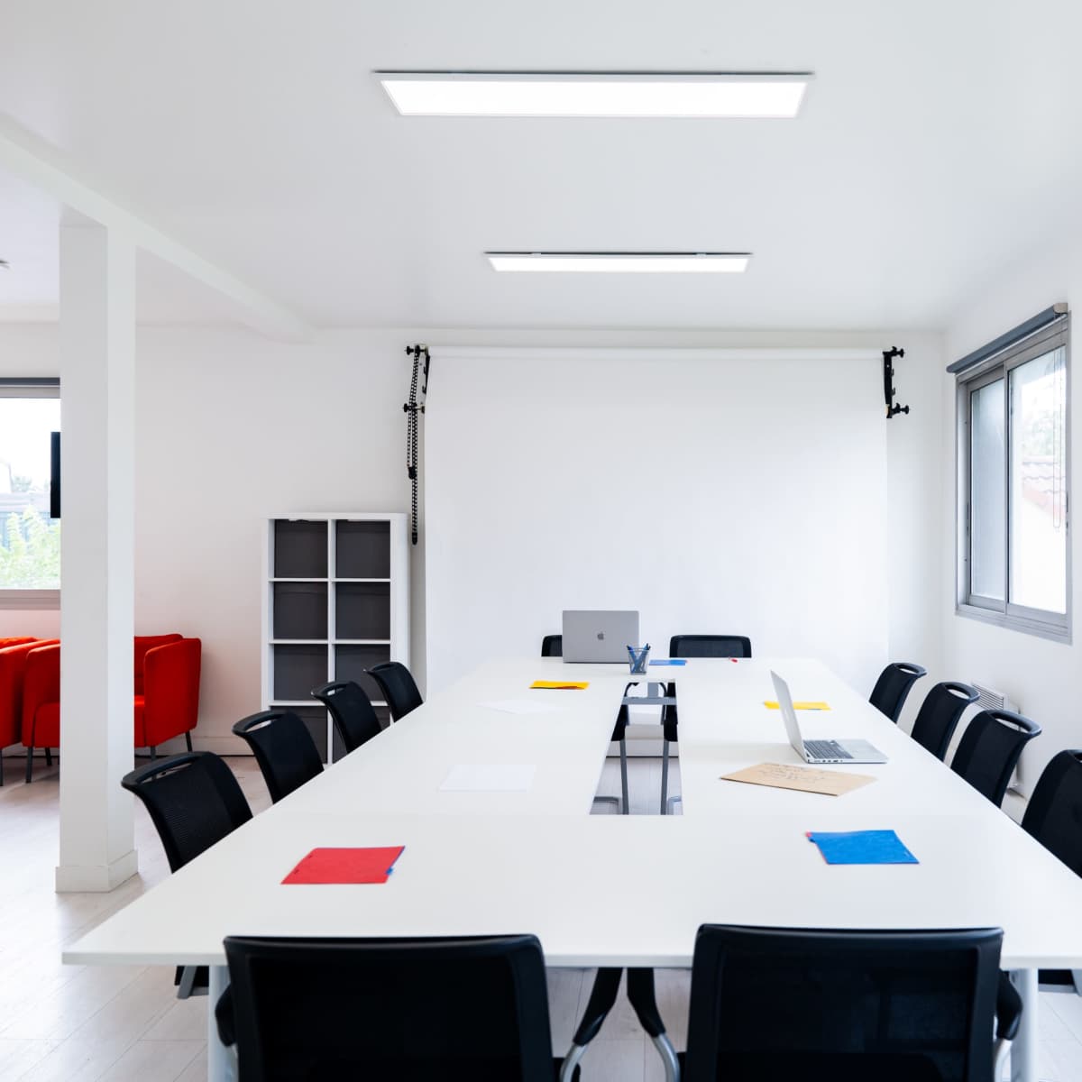 A large, modern meeting room features a long rectangular table with black chairs, a laptop, a monitor, and colored sheets of paper, all under bright overhead lighting and surrounded by windows.