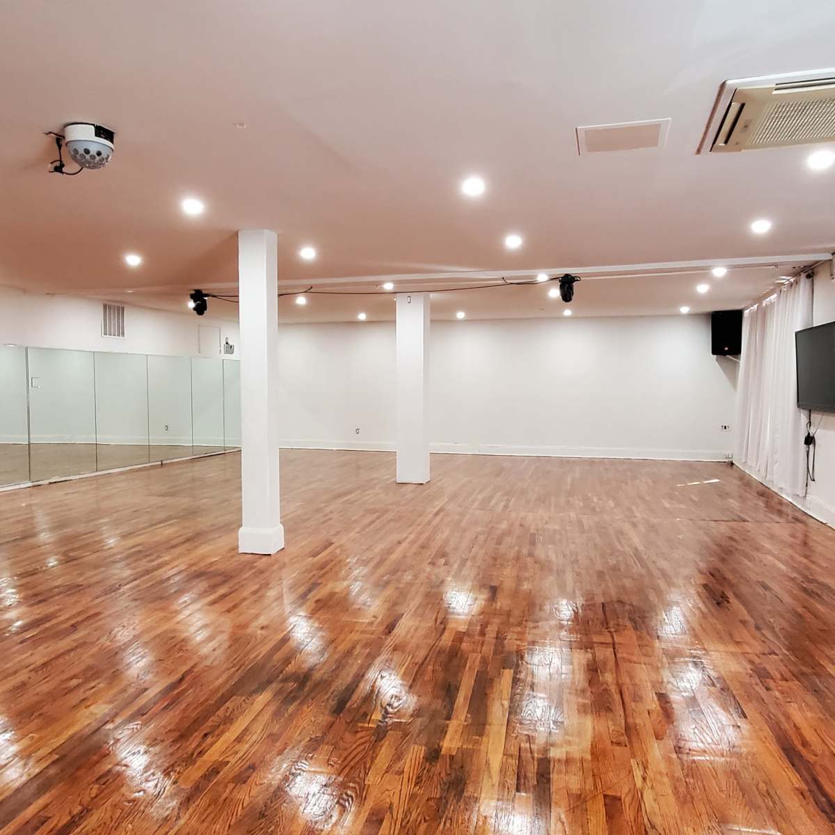 A spacious dance studio with polished wooden floors, tall mirrors on one wall, and white drapes covering large windows.