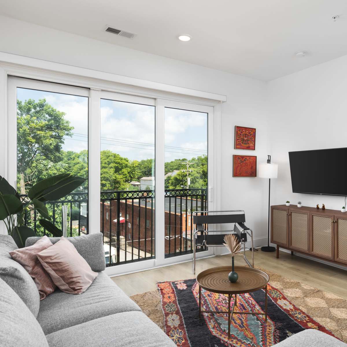 A modern living room features a gray sofa, a coffee table, a television mounted on the wall, and large windows that overlook a balcony filled with greenery.