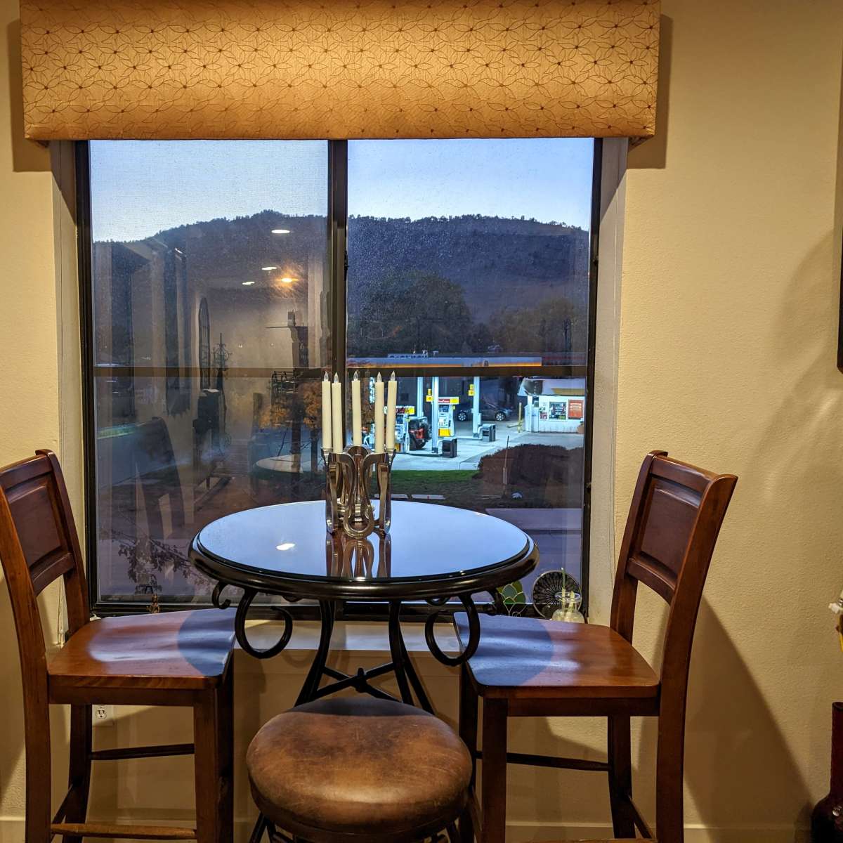 A small round table with two chairs is positioned near a window that overlooks a gas station and mountains in the background.