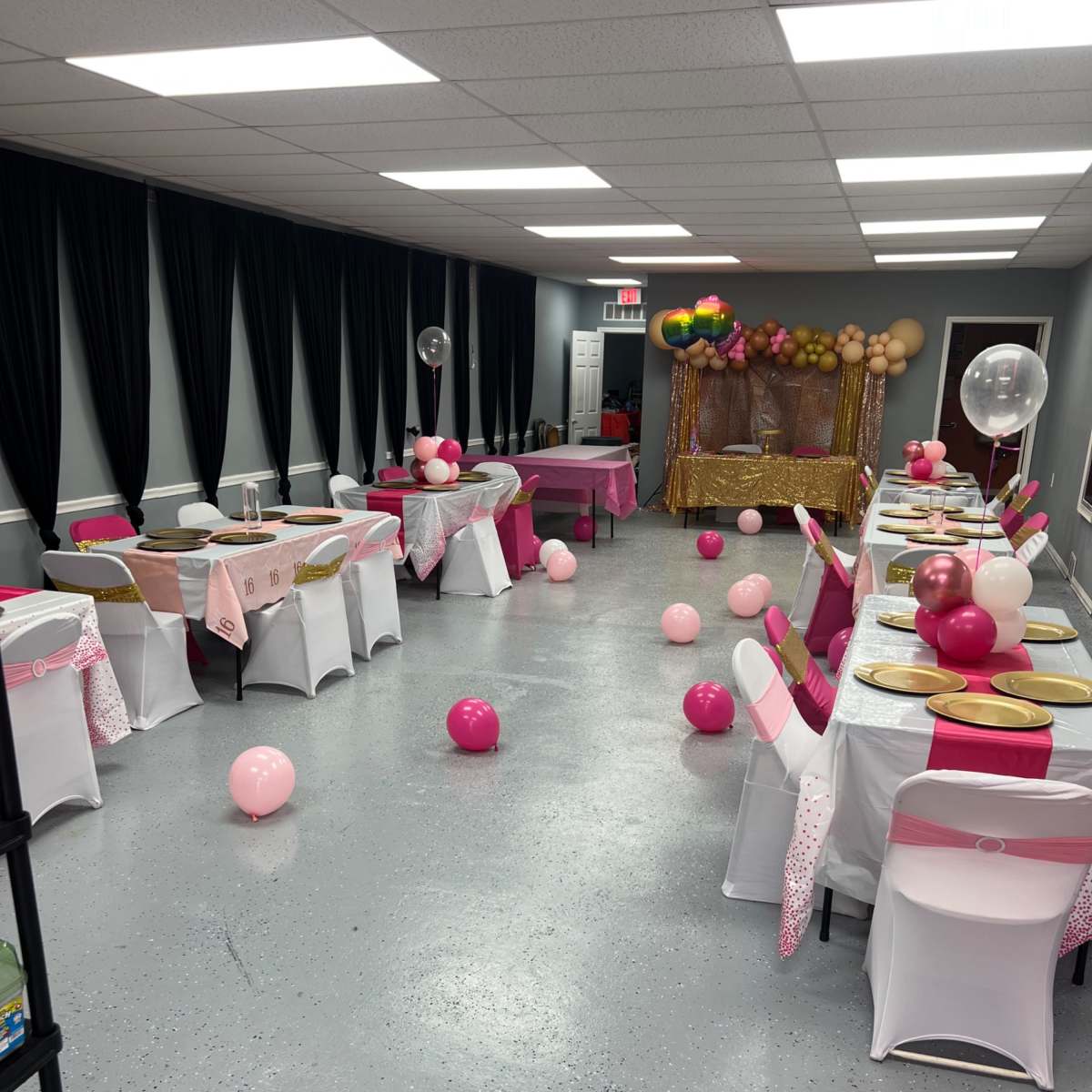 A decorated party space with tables draped in white and pink tablecloths, scattered balloons, and a backdrop featuring gold and pink decorations.