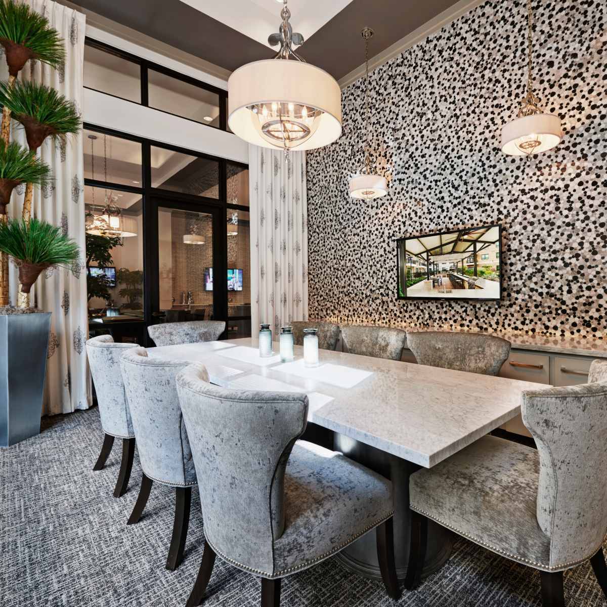 A modern dining area features a large marble table surrounded by upholstered chairs, with a textured wall adorned in black and white tiles and a tall potted plant in the corner.