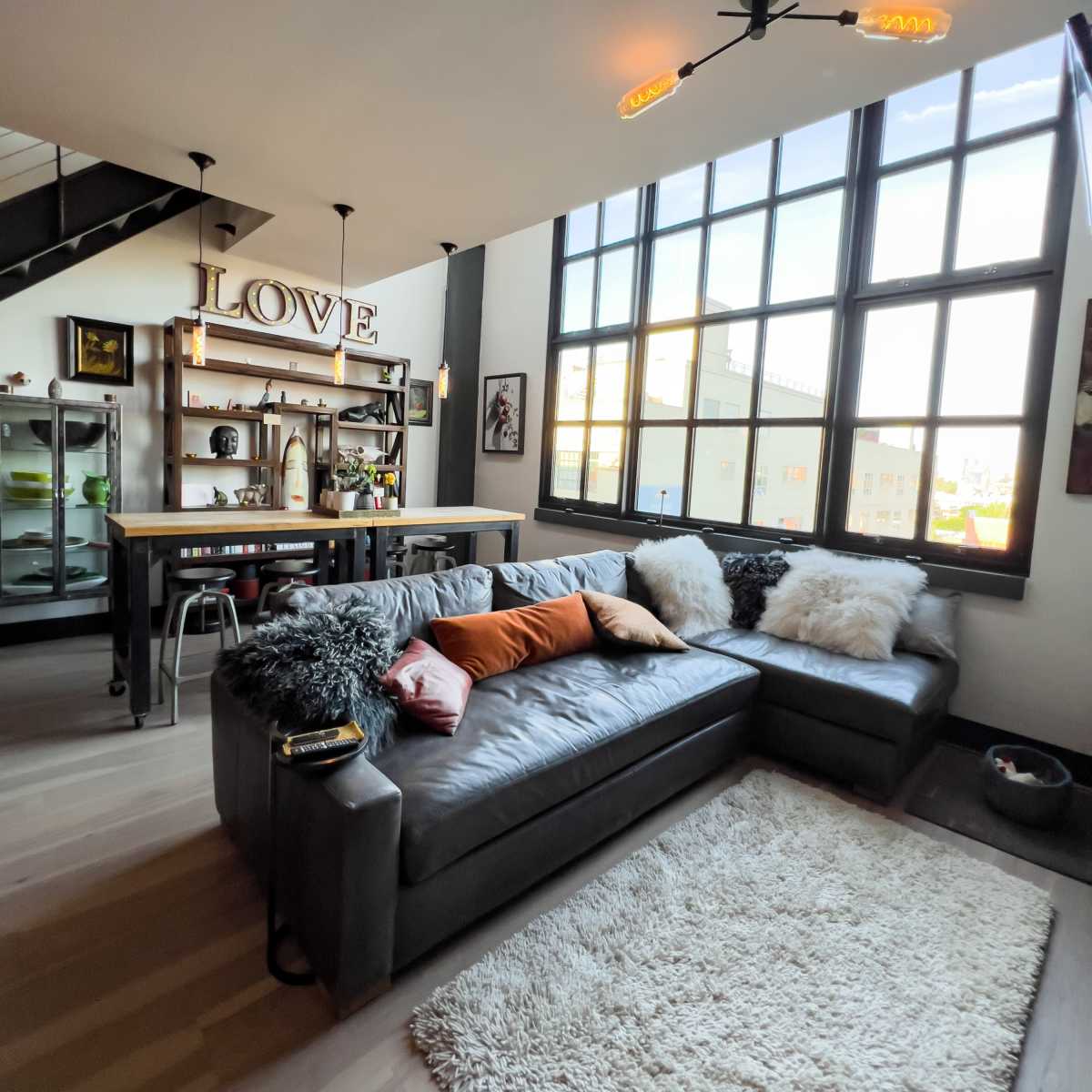 A modern living room featuring a large gray sectional sofa, a decorative rug, an industrial-style bar area, and large windows allowing natural light to enter the space.