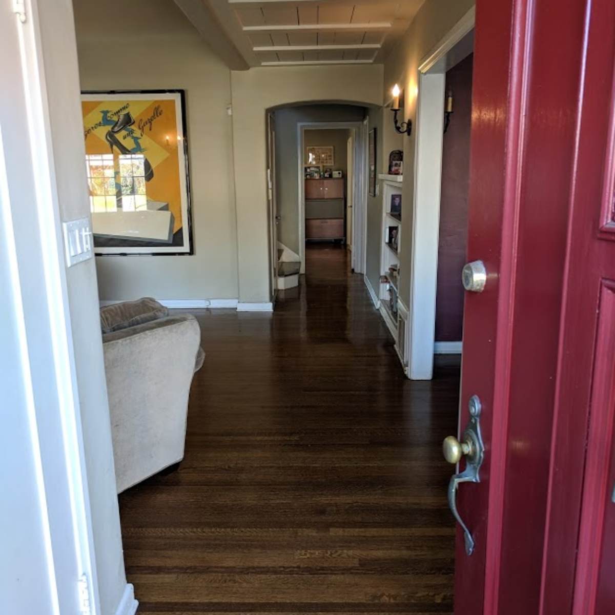 A view of a hallway from an open red front door, leading to a living area with wooden floors and white walls.