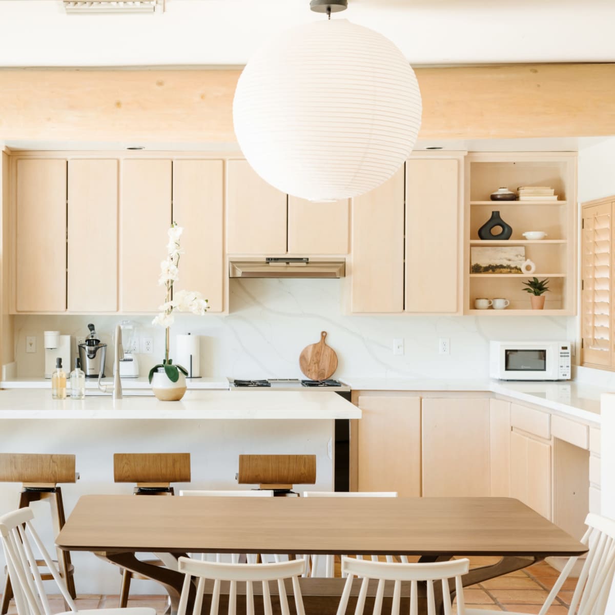 A modern kitchen features light wood cabinetry, a central island, and a dining table surrounded by white chairs.