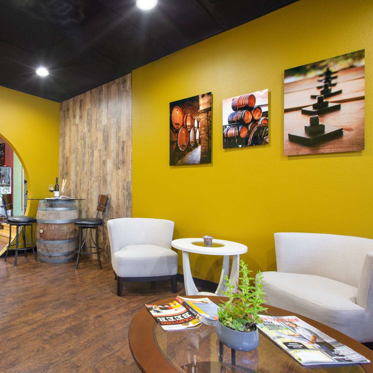 A cozy lounge area with two white chairs, a red chair, a round table, and a bar in the background, featuring wood accents and framed photographs on the walls.