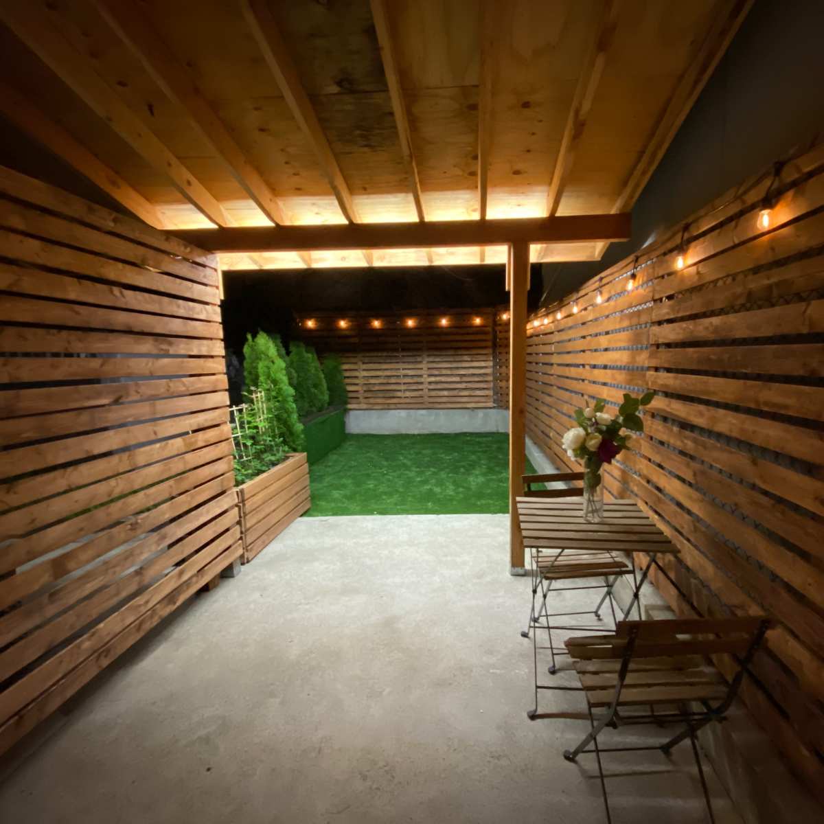 A secluded outdoor space with wooden slatted walls, artificial grass, and two metal chairs set beside a small table under warm string lights.
