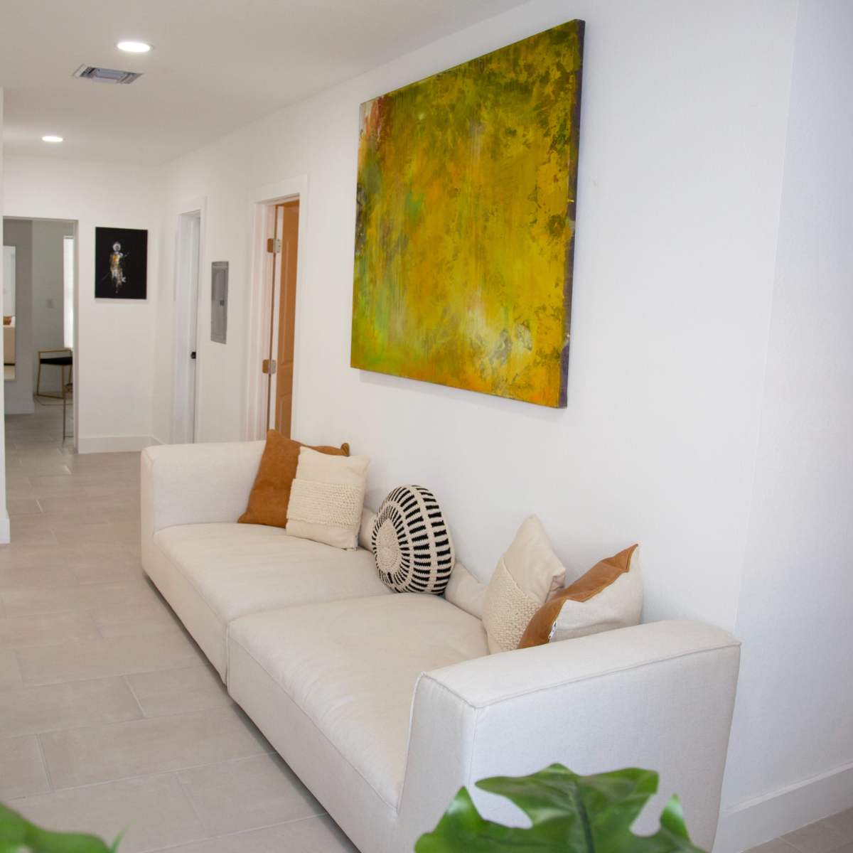 A hallway features a white sofa adorned with pillows and a large abstract painting on the wall, with a view into another room visible at the far end.