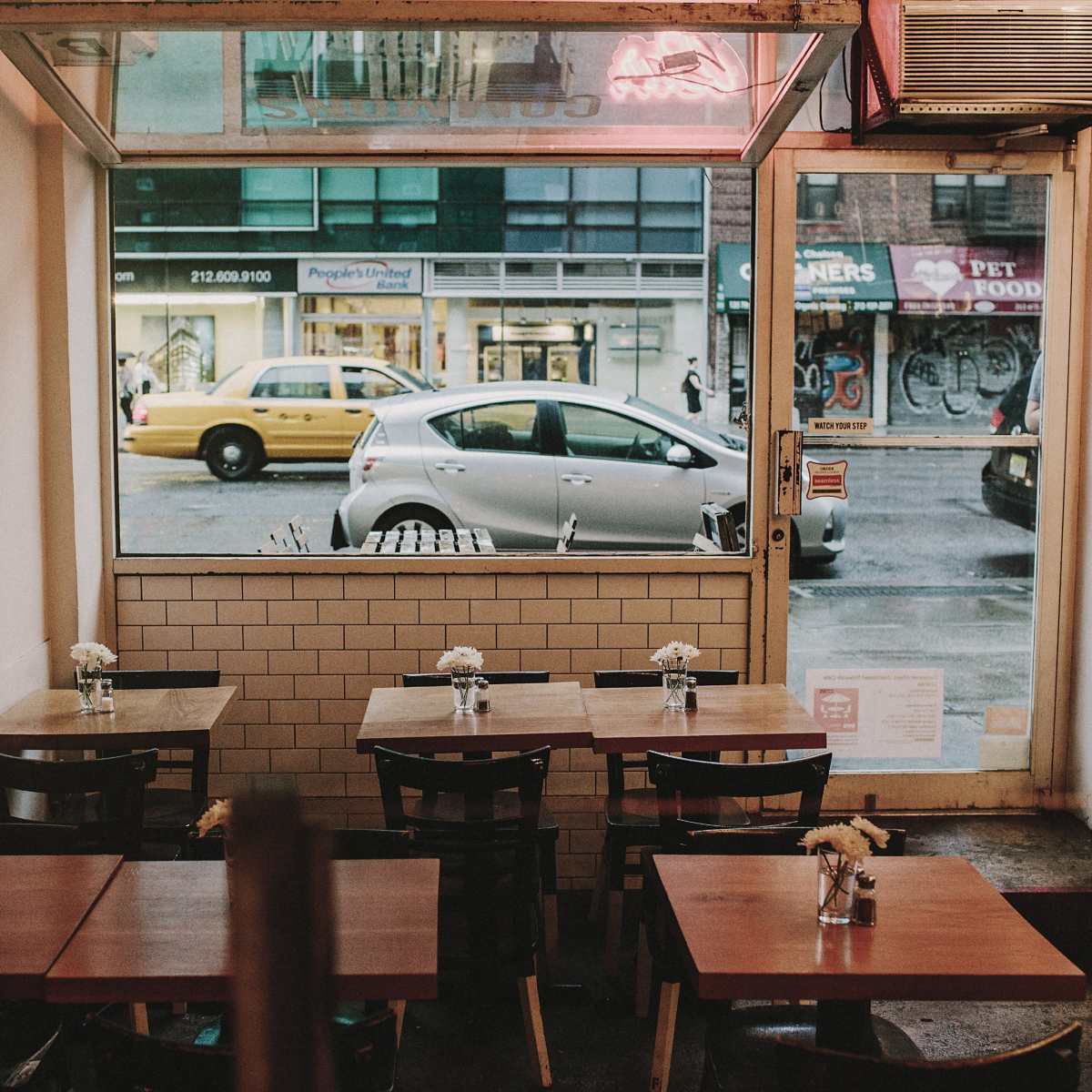 A small café with empty wooden tables and a view of a street with a parked car and a yellow taxi outside.