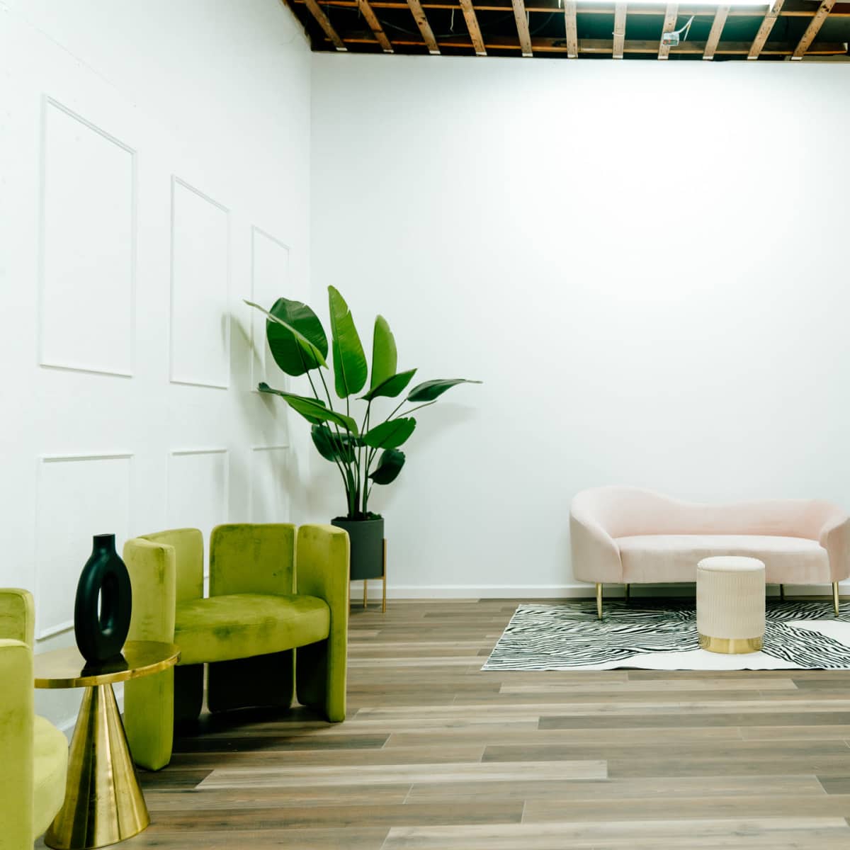 A minimalist room with a pink sofa, green chairs, a circular stool, a large mirror, and decorative plants against a light-colored wall.