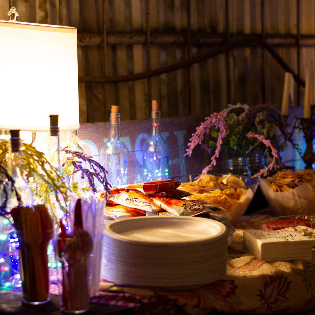 A table set up with various snacks and drinks, illuminated by lamps and decorated with colorful lights and flowers.