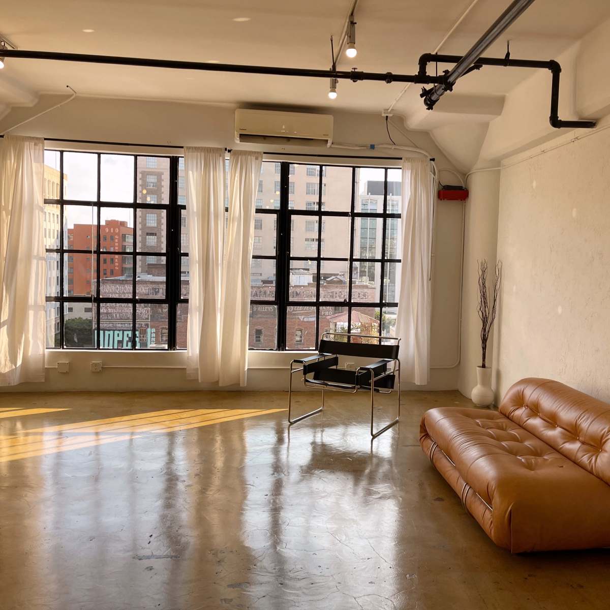 A spacious, well-lit room with large windows, a brown couch, and a black chair, featuring a concrete floor and light-colored walls.